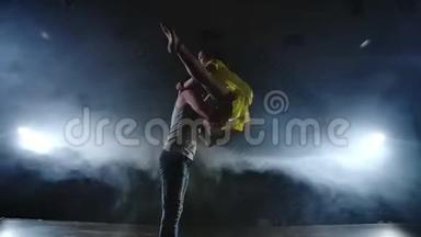 两名现代芭蕾舞演员正在舞台上用烟熏火燎的方式跳舞，一名男子举起他的舞伴，旋<strong>转进</strong>来